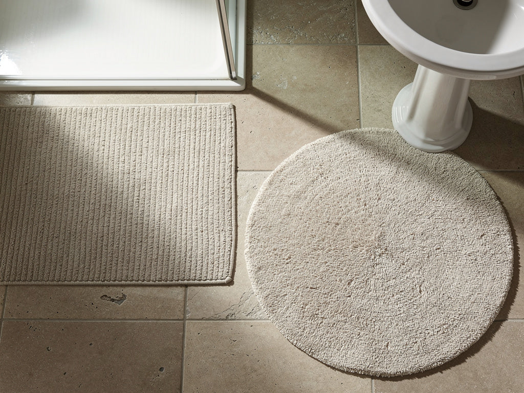 A ribbed bath mat beside a shower and a round bath rug in front of a sink, shown on the same bathroom floor.