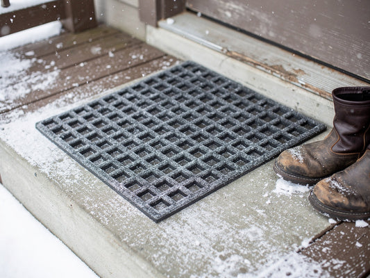 A charcoal rubber-backed doormat on a snow-dusted porch step next to a pair of snow boots.