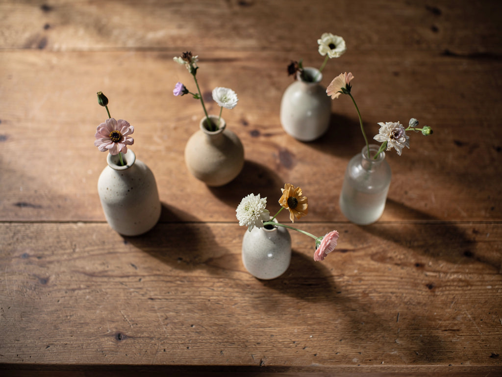 Five small bud vases on a wood table, each holding a single flower stem.