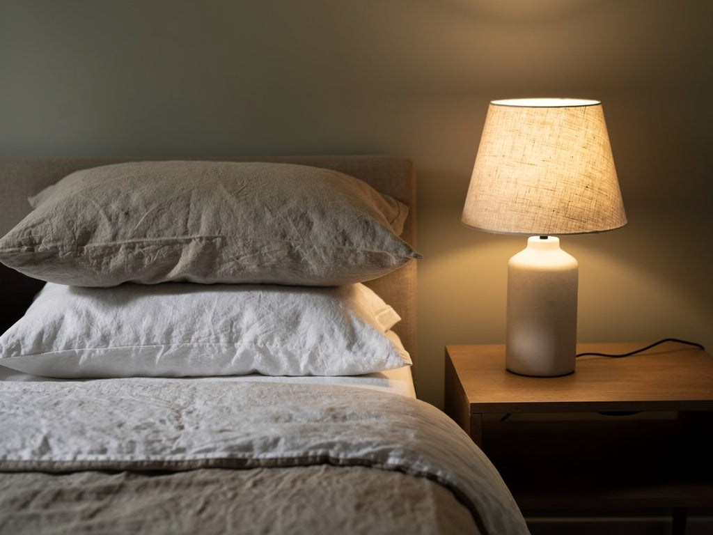A lit bedside table lamp on a nightstand next to a bed with two pillows, showing the lamp's height relative to the bed.