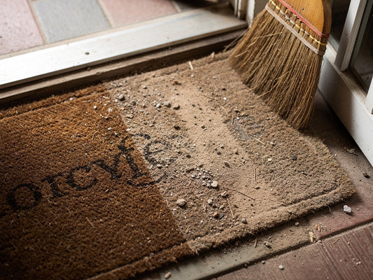 A doormat on a front step, partly cleaned, with a broom leaning beside it
