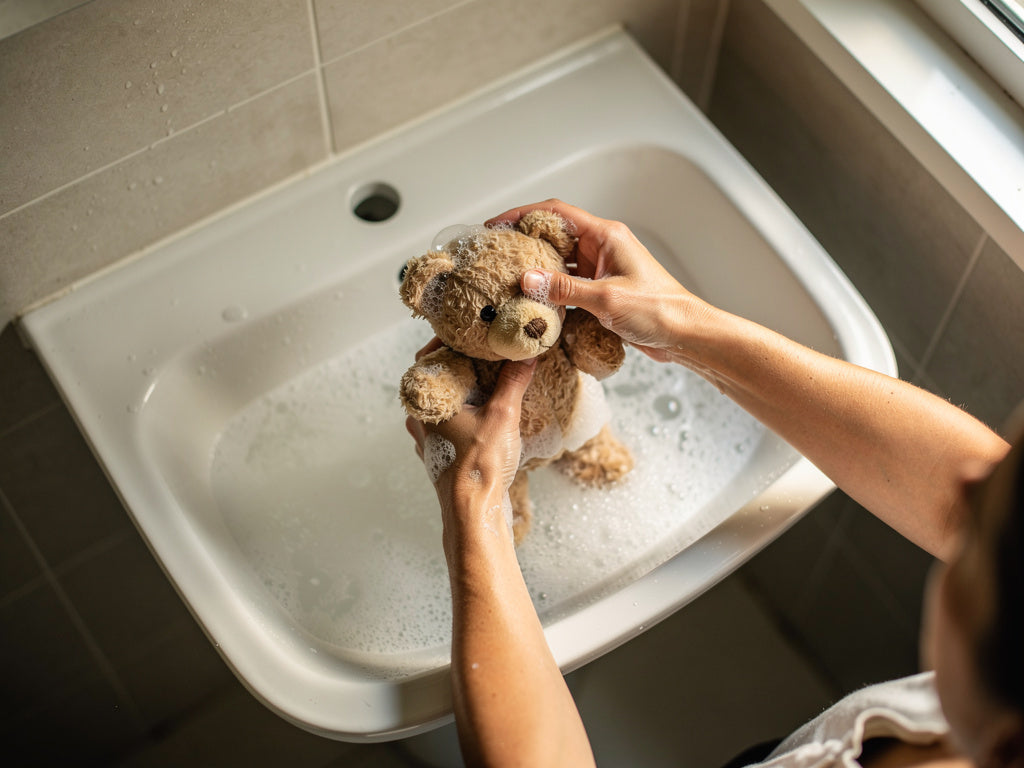 Hands washing a teddy bear by hand in a sink full of soapy water.