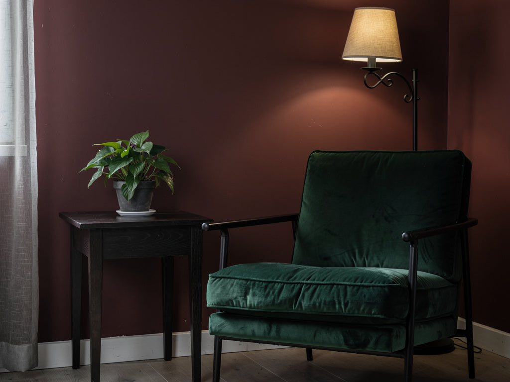 A living room corner decorated in a gothic style with an oxblood accent wall, a velvet armchair, and a wrought iron floor lamp.