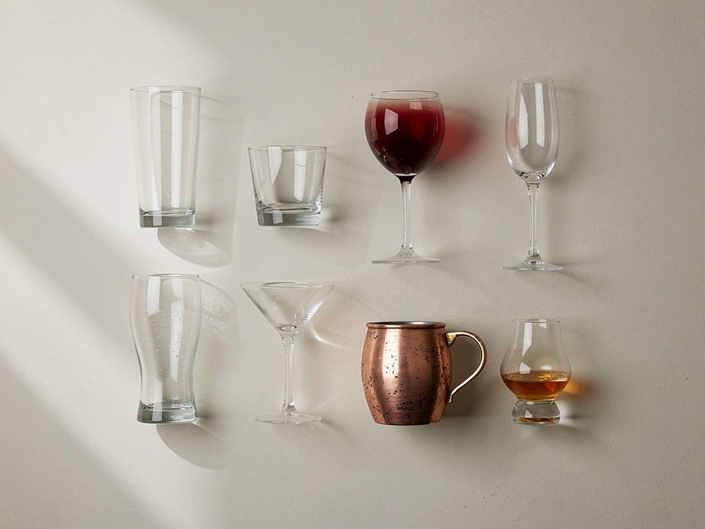 A grid of eight different drinking glass shapes, including a highball, a rocks glass, a wine glass, a champagne flute, a martini glass, a pilsner glass, a copper mug, and a Glencairn glass, arranged for comparison.