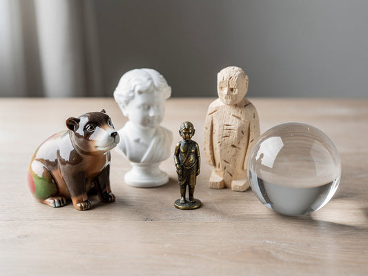 Five decorative figurines in resin, porcelain, metal, wood, and glass grouped together.