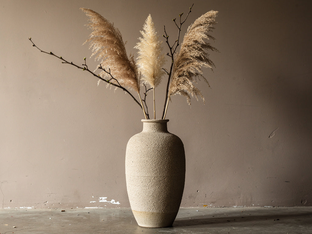 Tall floor vase filled with dried pampas grass and branches against a plain wall.
