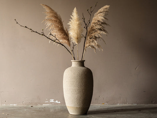 Tall floor vase filled with dried pampas grass and branches against a plain wall.