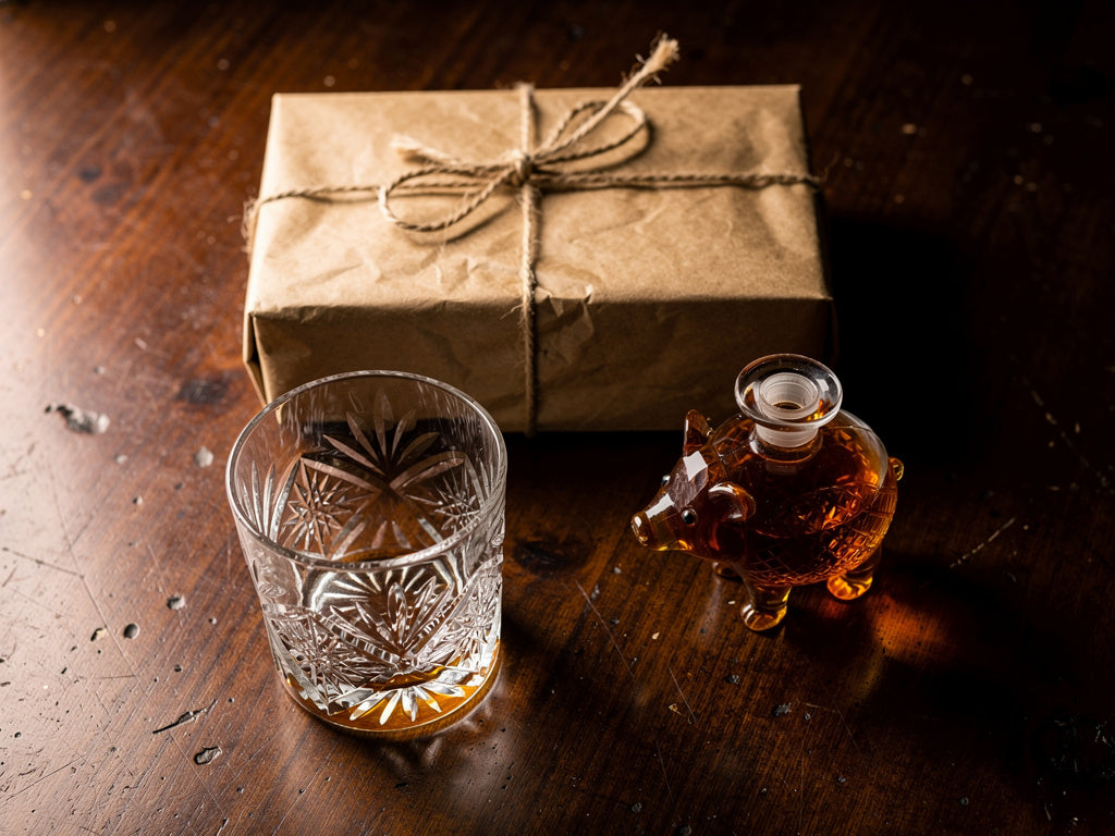 A hand-cut whiskey glass, a small decanter, and a wrapped gift box arranged on a dark wood table.