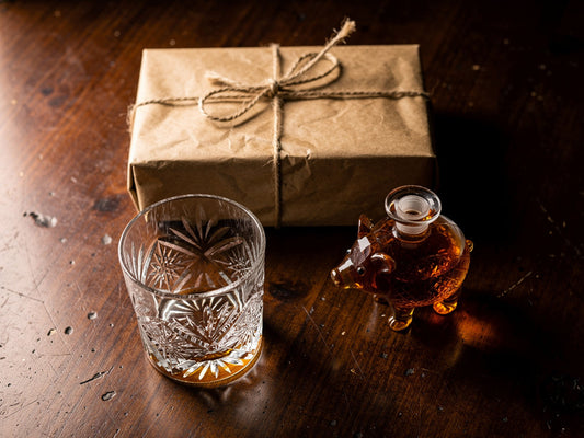 A hand-cut whiskey glass, a small decanter, and a wrapped gift box arranged on a dark wood table.